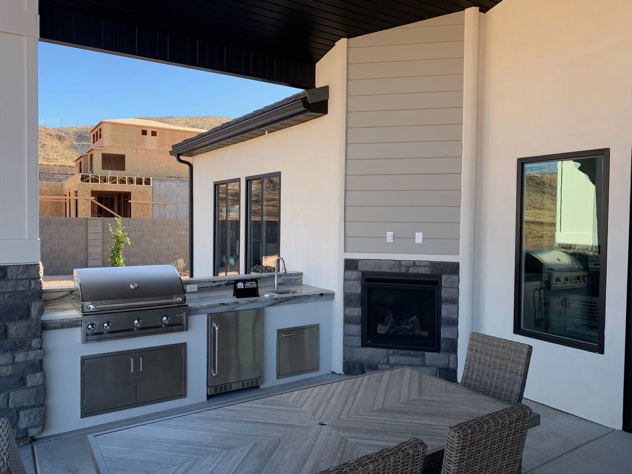 Covered patio outdoor kitchen with a white stucco island, built-in stainless grill, stone countertops, sink, outdoor refrigerator, and a stone-surround gas fireplace in St. George, Utah