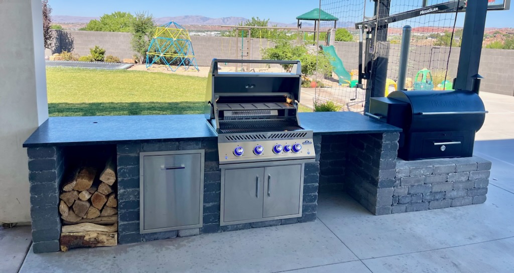 Stacked stone outdoor kitchen with a Napoleon built-in grill, dark stone countertop, stainless steel doors, built-in firewood storage, and a raised stone smoker platform in St. George, Utah